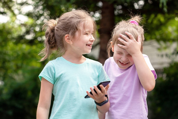 Two elementary school age children, girls, sisters siblings laughing, having fun using a smartphone, mobile phone, outdoors portrait, tech savvy kids and technology concept, real people lifestyle - Foto, Imagem