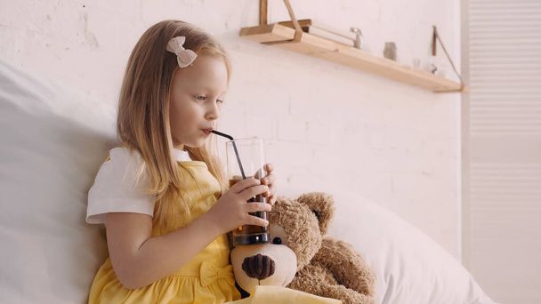 Girl drinking juice with straw near teddy bear on bed  - Foto, afbeelding