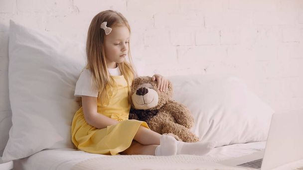 Enfant regardant l'ordinateur portable flou près de l'ours en peluche sur le lit  - Photo, image