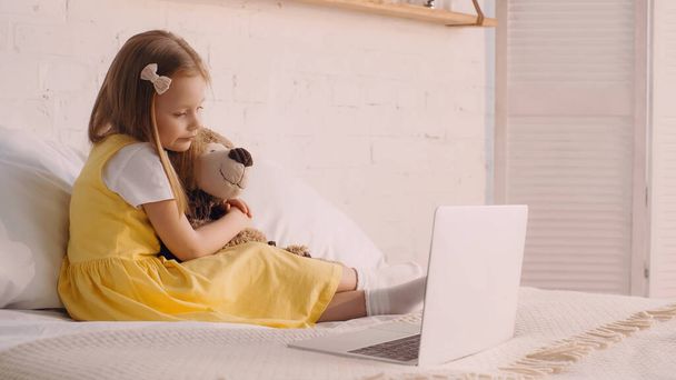 Preschooler child hugging soft toy near laptop on bed at home  - Photo, Image