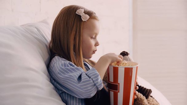 Side view of kid holding popcorn near soft toy in bedroom  - Foto, Bild