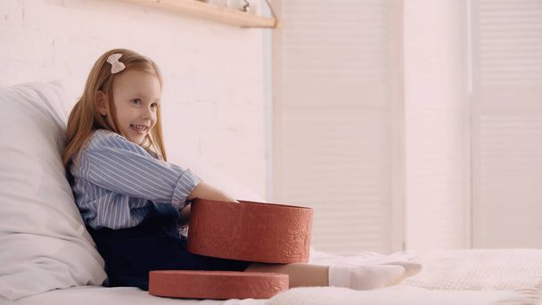 Cheerful kid holding gift box on bed at home  - Foto, afbeelding