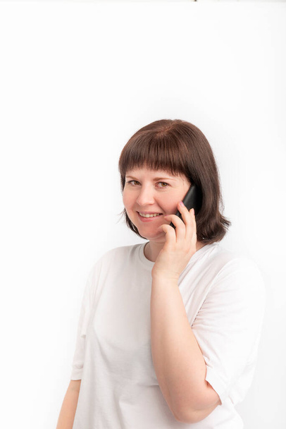 Una chica sonriente hablando por teléfono. Retrato de una linda mujer de cara redonda con un teléfono en sus manos sobre fondo blanco - Foto, imagen