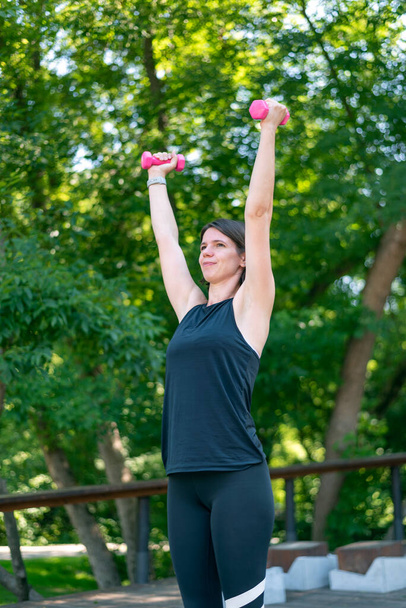 Fiatal sportoló nő fitnesz súlyzókkal a szabadban. A lány feltartott kézzel tartja a súlyzókat a zöld lombozat hátterében. - Fotó, kép