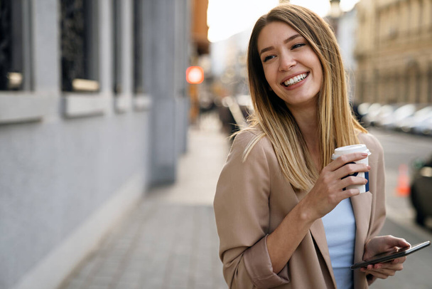 Portrait of happy successful business woman with coffee and digital tablet on her way to work on city street. - Zdjęcie, obraz