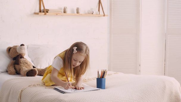 Child drawing on paper near color pencils and soft toy on bed  - 写真・画像