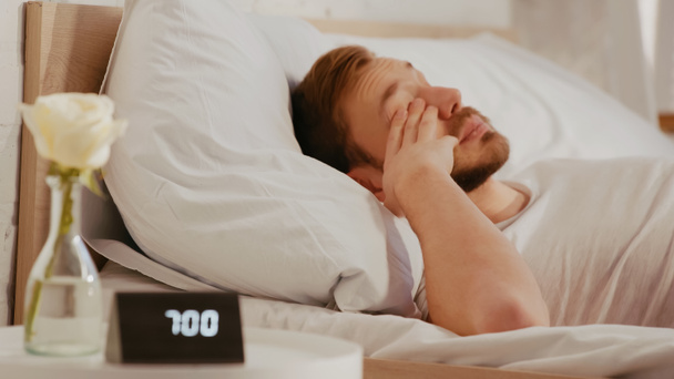 Sleepy man lying on bed near blurred clock and flower in bedroom  - Foto, afbeelding