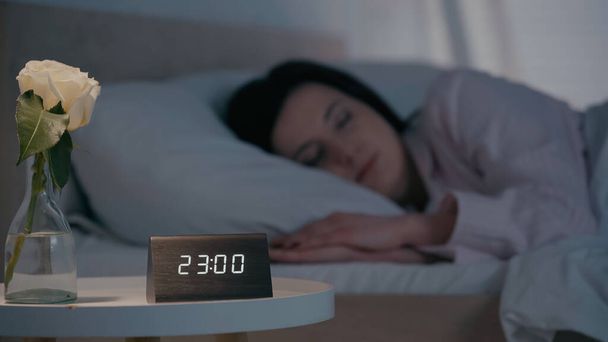 Clock and flower on bedside table near blurred woman sleeping on bed at night  - Фото, изображение