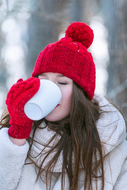 Beautiful brunette girl in red hat and mittens drinks tea outdoors. Vertical frame. - Foto, afbeelding