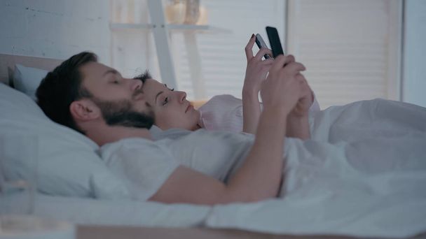 Young woman using smartphone near husband on bed at home  - Φωτογραφία, εικόνα