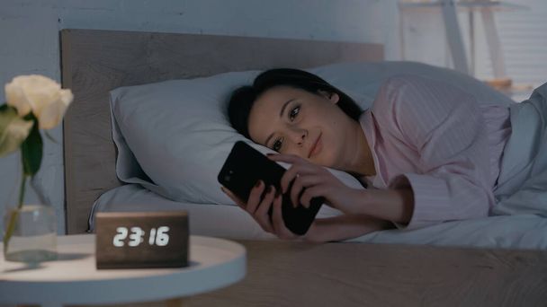 Brunette woman using smartphone on bed near blurred clock in bedroom  - Photo, Image