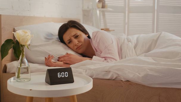 Upset woman looking at clock on bedside table in bedroom  - Zdjęcie, obraz
