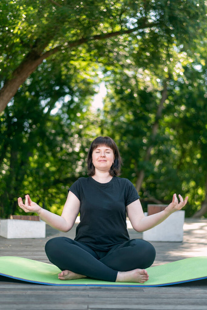 Güzel genç kız parkta meditasyon yapıyor. Lotus pozisyonunda yeşil bir yoga minderinin üzerinde oturuyor. Sağlıklı bir yaşam tarzı. Dikey çerçeve. - Fotoğraf, Görsel