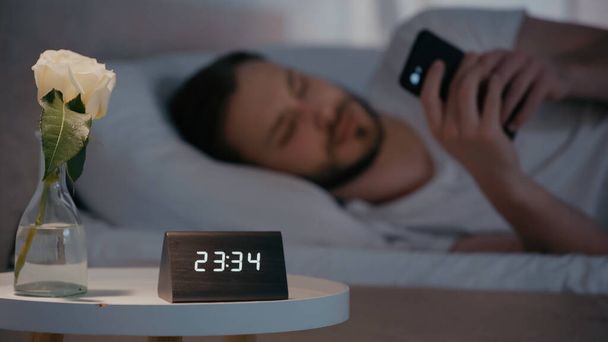 Plant and clock on bed near blurred man using smartphone on bed at home  - Foto, Imagem