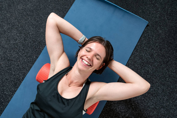 Портрет молодой улыбающейся спортивной женщины, лежащей на миофасциальном рулоне. Миофасциальный выпуск с розочкой. Вид сверху. - Фото, изображение