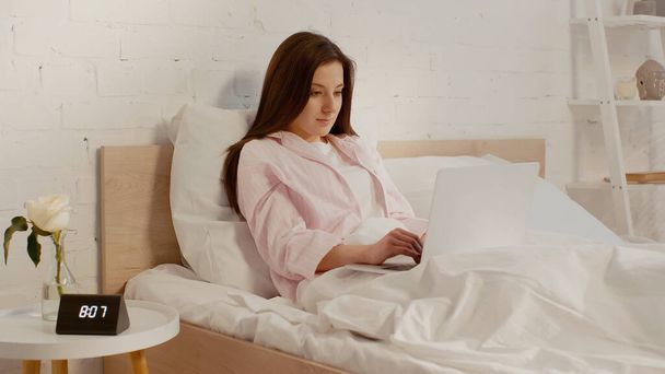 Nuori brunette nainen käyttää kannettavaa tietokonetta sängyssä lähellä kasvi ja kello yöpöydällä  - Valokuva, kuva