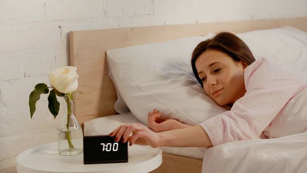Young woman turning off clock near flower in bedroom  - Fotó, kép