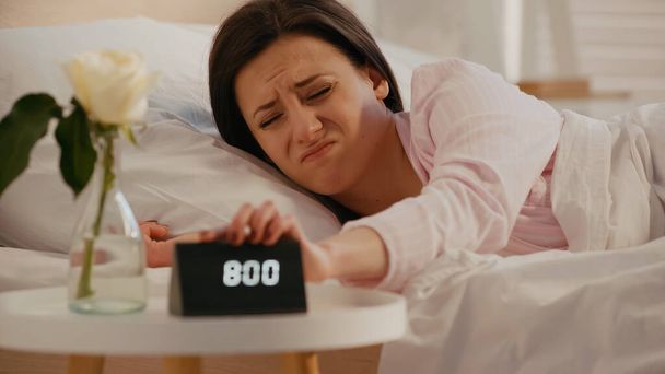 Displeased woman turning off clock near blurred plant in bedroom  - Фото, изображение