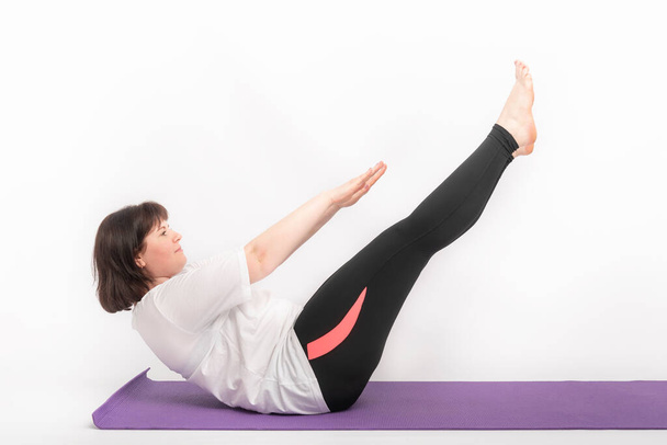 Overgewicht vrouw sport op yoga mat. Meisje rockt met de buikspieren. Zijaanzicht op witte achtergrond - Foto, afbeelding
