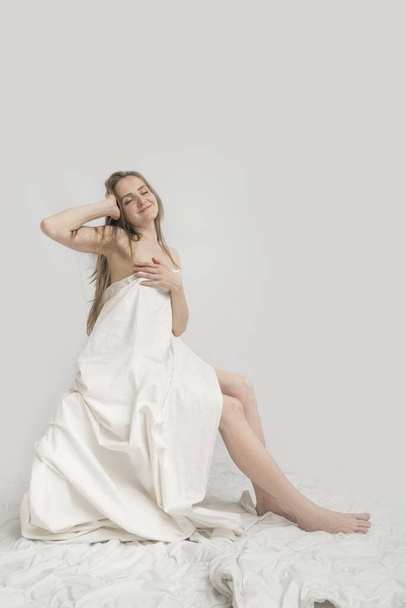 Young caucasian female model is sitting covered herself with white sheet and poses. Concept of good morning. White background. Vertical frame. - Фото, изображение