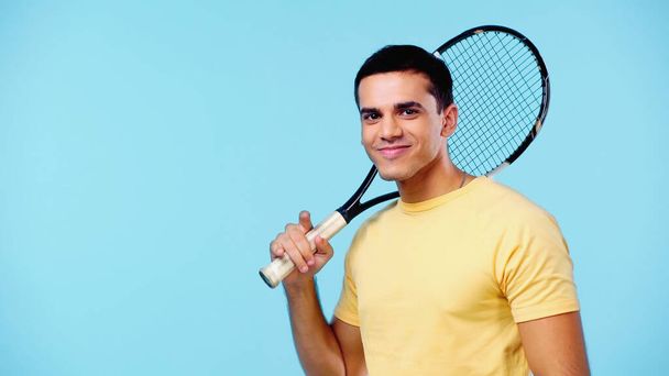 šťastný mladý muž ve žlutém tričku drží tenisovou raketu izolované na modré - Fotografie, Obrázek