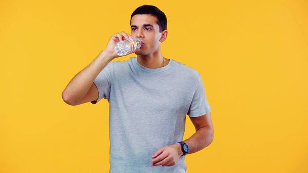 young man in t-shirt holding bottle while drinking water isolated on yellow - Foto, Imagen