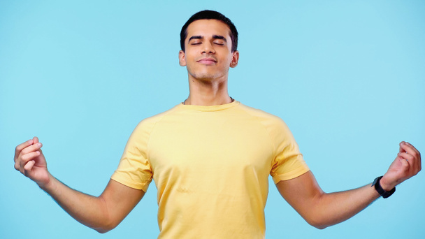 pleased young man with closed eyes in yellow t-shirt meditating isolated on blue - Valokuva, kuva