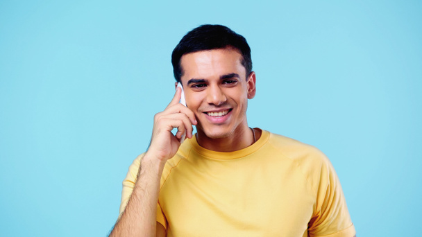 joven positivo en camiseta amarilla hablando en el teléfono celular aislado en azul - Foto, Imagen