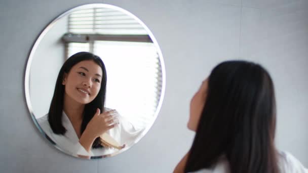 Mujer coreana feliz cepillando el pelo mirando el espejo en el baño - Metraje, vídeo