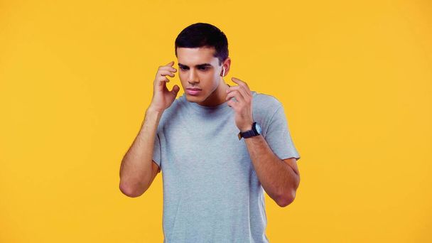 confused young man in t-shirt adjusting wireless earphones isolated on yellow  - Photo, Image