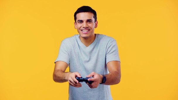 KYIV, UKRAINE - DECEMBER 22, 2021: joyful young man holding joystick and playing video game isolated on yellow - Fotó, kép