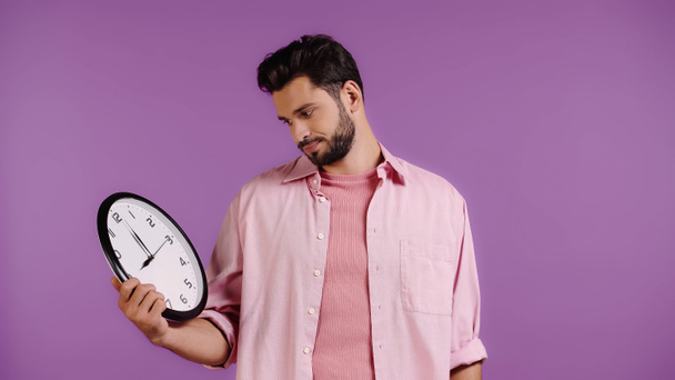 nespokojený mladý muž se dívá na hodiny izolované na fialové  - Fotografie, Obrázek