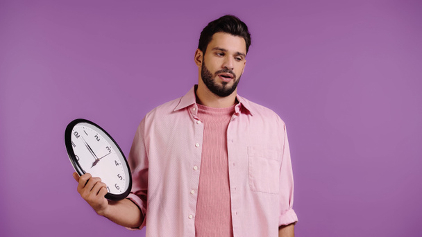 Unzufriedener junger Mann hält Uhr isoliert auf lila  - Foto, Bild