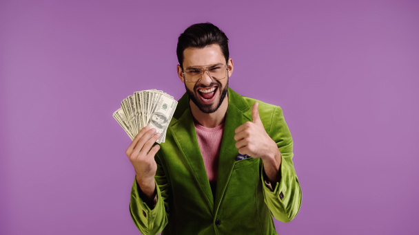 Χαρούμενος άντρας με πράσινο μπουφάν και γυαλιά που κρατάει δολάρια και δείχνει τον αντίχειρα του απομονωμένο στο μωβ. - Φωτογραφία, εικόνα