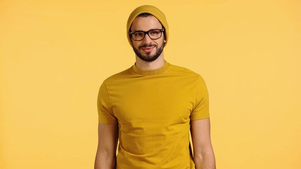 Ευτυχισμένος νεαρός άνδρας με καπέλο, γυαλιά και t-shirt που απομονώνονται σε κίτρινο - Φωτογραφία, εικόνα