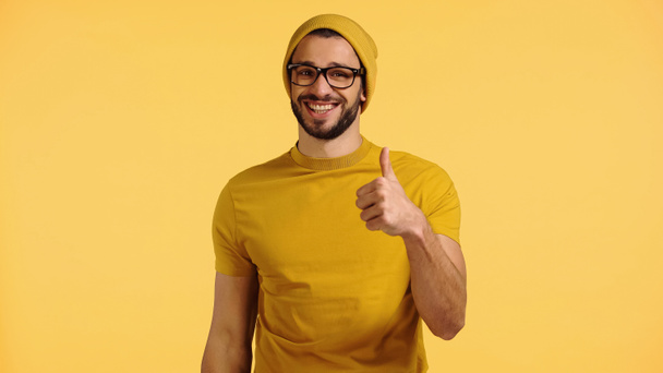 happy young man in beanie hat, glasses and t-shirt showing thumb up isolated on yellow - Foto, Bild