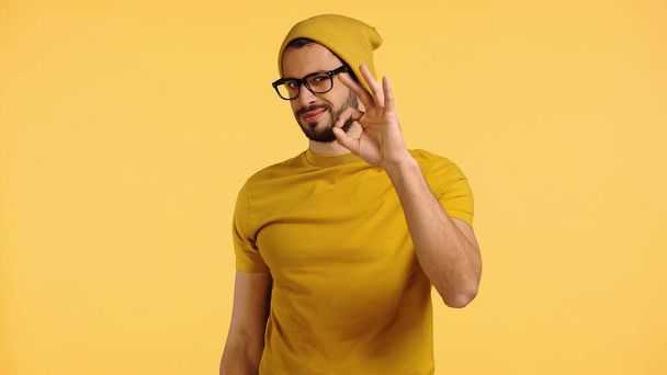 young man in beanie hat, glasses and t-shirt showing okay isolated on yellow - Fotó, kép