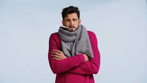 sick man in pink sweater and scarf standing with crossed arms while measuring temperature isolated on blue  - Zdjęcie, obraz