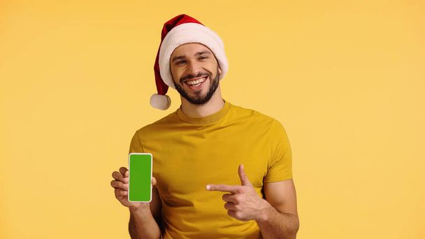 サンタ・ハットの幸せな男は指で指差す黄色に孤立したスマートフォンで - 写真・画像