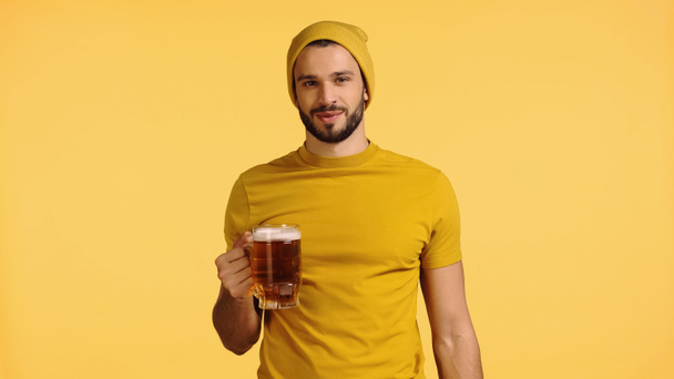 Bere şapkalı ve tişörtlü genç adam elinde cam bardakla sarıya izole edilmiş bira tutuyor. - Fotoğraf, Görsel