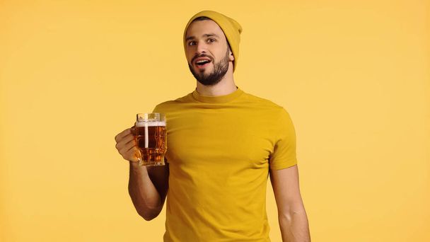 amazed man in beanie hat and t-shirt holding glass mug with beer isolated on yellow - Фото, изображение