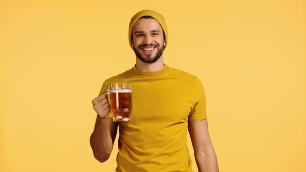 hombre feliz en gorro sombrero y camiseta sosteniendo taza de vidrio con cerveza aislada en amarillo - Foto, Imagen