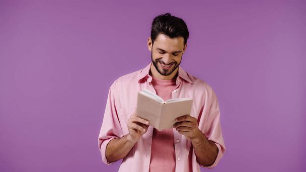 cheerful bearded man reading book isolated on purple - Foto, afbeelding