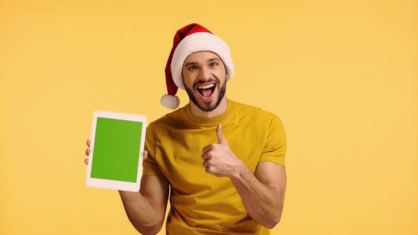 felice uomo in cappello di Babbo Natale tenendo tablet digitale e mostrando pollice in su isolato su giallo - Foto, immagini