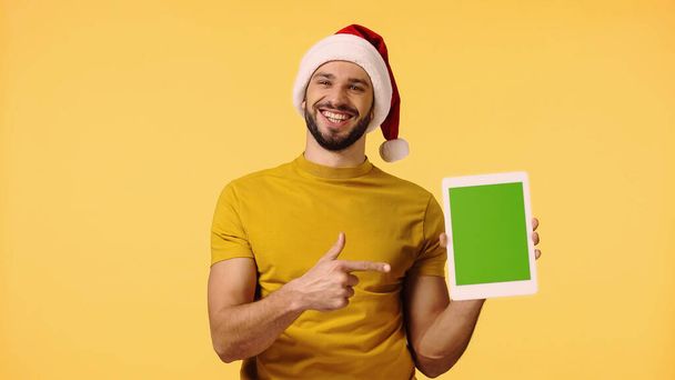 šťastný muž v Santa klobouk ukazuje prstem na digitální tablet izolované na žluté - Fotografie, Obrázek