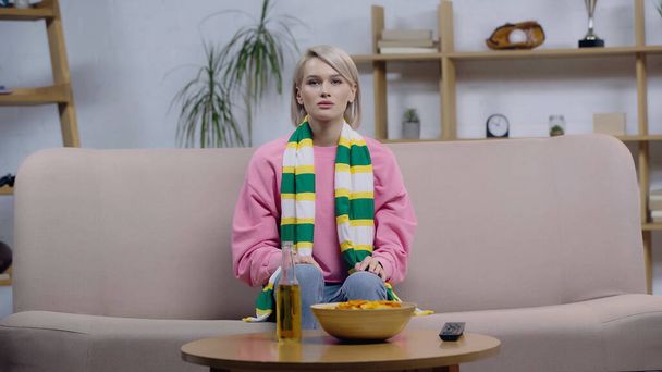 frustrated sport fan woman in striped scarf watching match on tv near chips and bottle of beer - Photo, Image