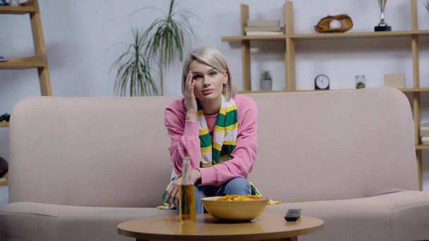 upset woman in striped scarf sitting on sofa near chips and beer while watching sport competition on tv - Photo, Image