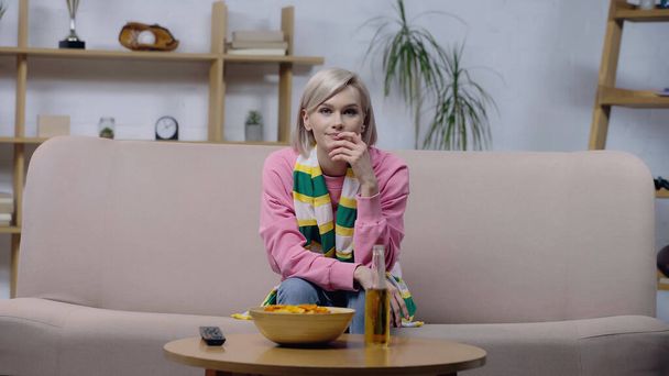 displeased woman in striped scarf watching sport championship on sofa near chips and beer - Foto, immagini