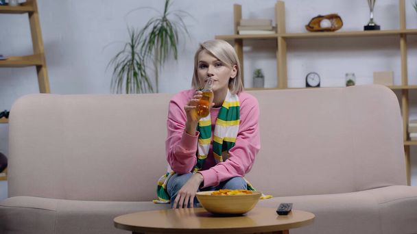 Блондинка в полосатом шарфе пьет пиво и смотрит чемпионат по телевизору дома - Фото, изображение