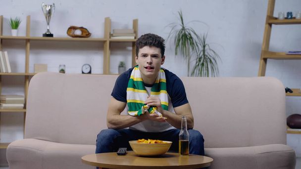 fanático del deporte emocionado sentado en el sofá cerca de las patatas fritas y la cerveza y viendo el juego en la televisión - Foto, Imagen